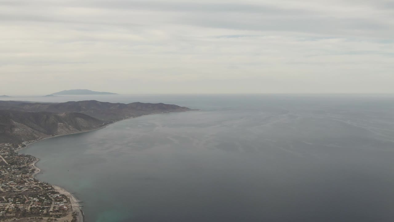 Aerial view of coastal homes, beach, ocean, mountains, and overcast sky.