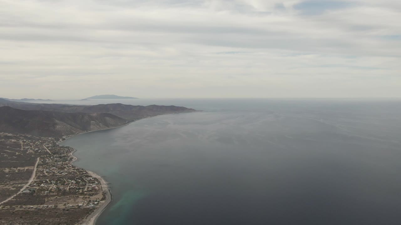 Wide aerial frame over Los Barriles, Baja California Sur, with the town and beach low left, broad blue-gray ocean center, distant mountains on the horizon, cloudy sky, and no visible people.