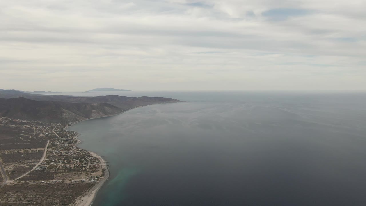 Aerial view of Los Barriles with more of the shoreline town visible at lower left, calm ocean filling the right side, low hills behind the coast, layered clouds, and no visible people.