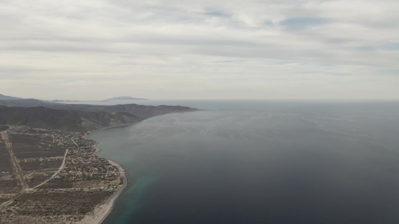 High drone frame over Los Barriles showing the beach curve at lower left, clustered buildings and roads inland, darker hills along the coast, expansive blue-gray water, cloudy sky, and no visible people.