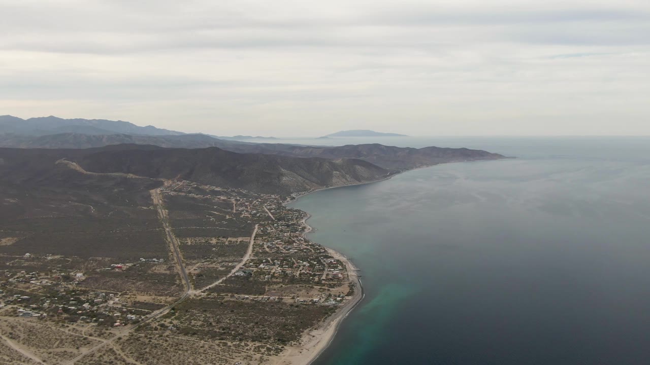 Aerial view of a coastal town, sandy beach, ocean, and hills under cloudy sky.