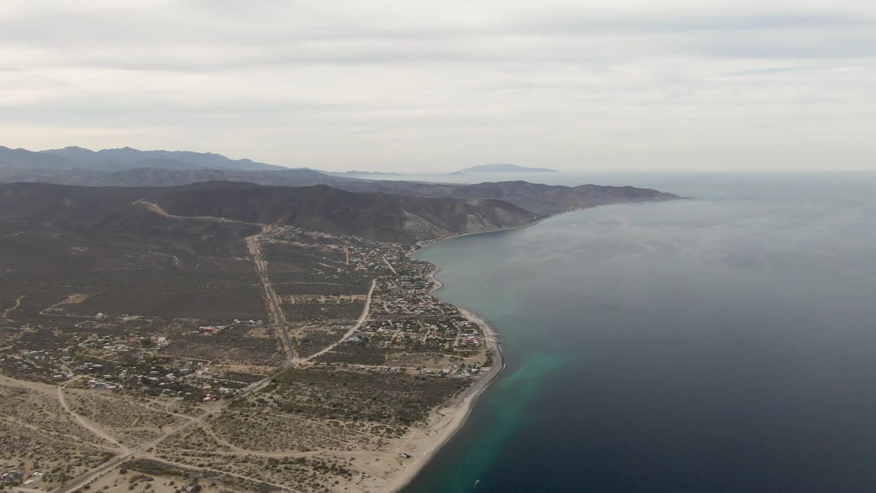 Aerial frame over Los Barriles, Mexico, with pale roads and scattered buildings in the foreground, curving sandy shoreline, blue water, hills, and no visible people.