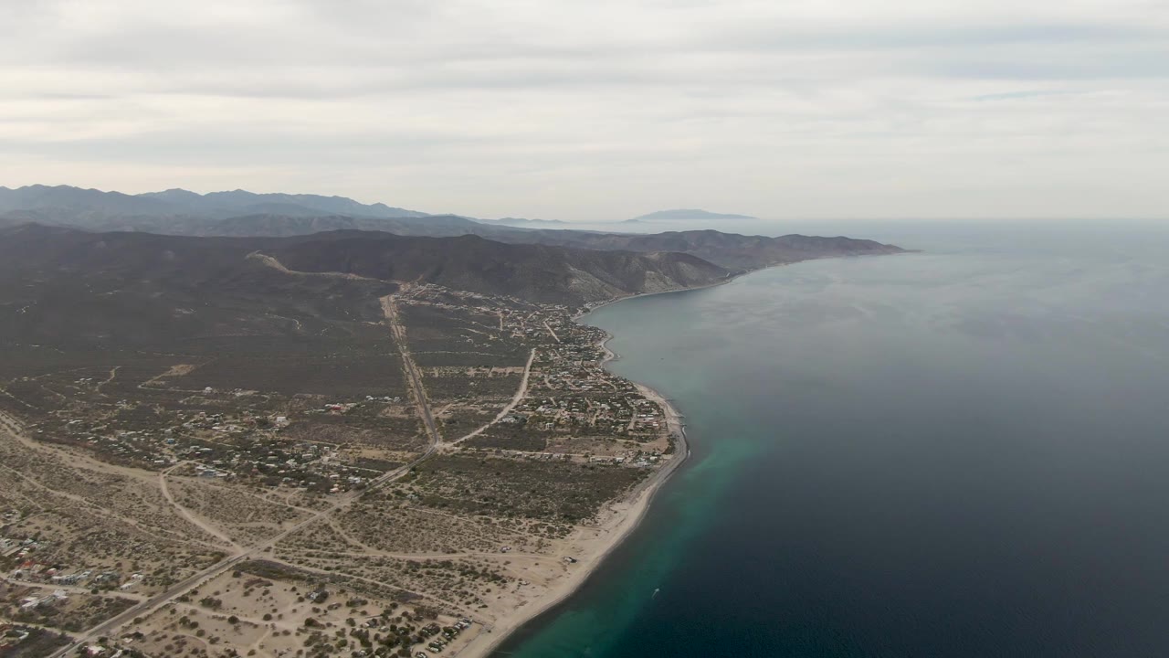 Aerial view in Los Barriles, Mexico, looking across desert roads and beach toward clear blue ocean, low coastal hills, a cloudy horizon, and no visible people.