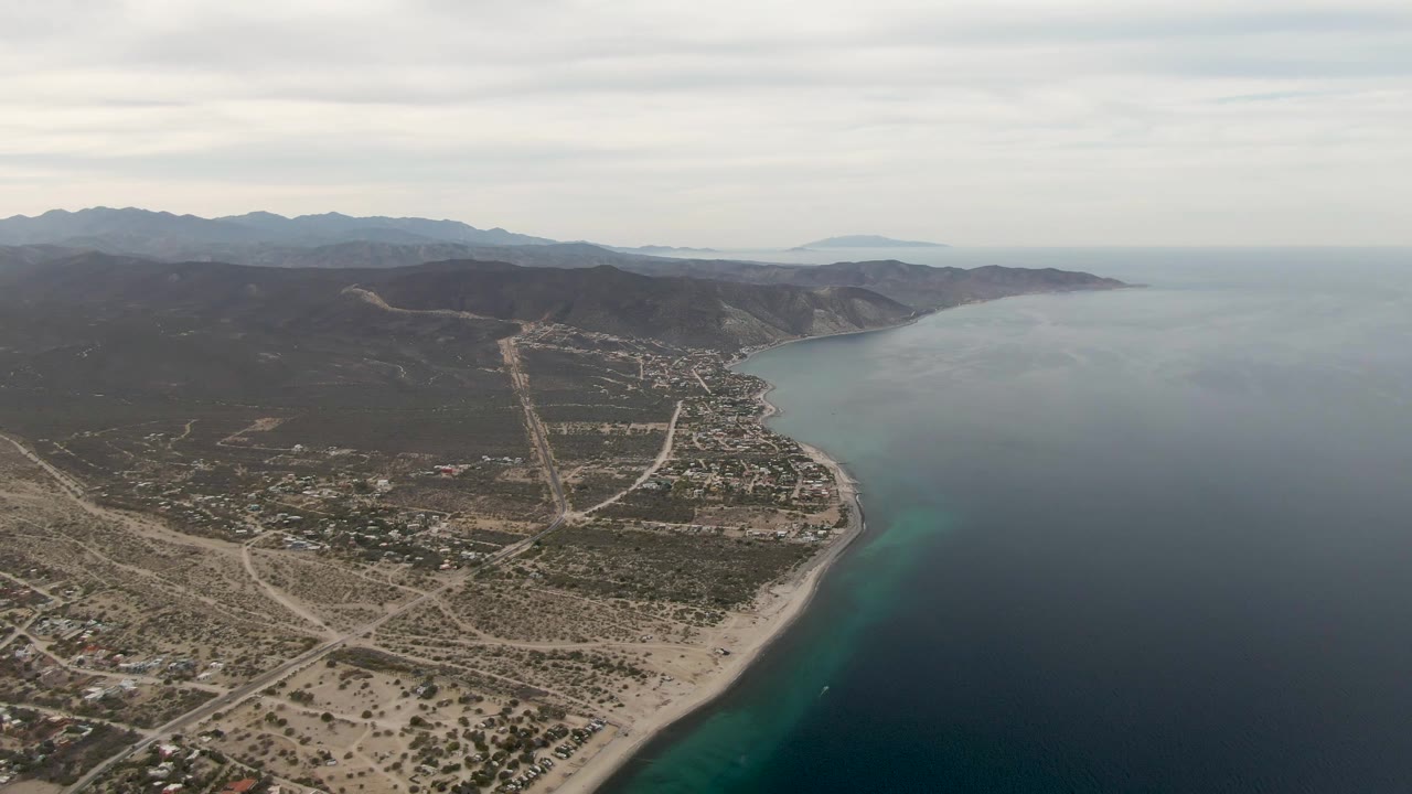 Mid-clip aerial frame over Los Barriles, Mexico, showing more of the shoreline curve, dense coastal buildings, pale roads, blue ocean, distant hills, and no visible people.