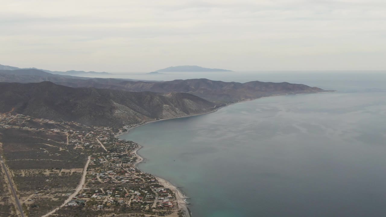 Aerial frame over Los Barriles, Baja California Sur, showing a coastal town at lower left, a curved bay, brown green mountains, hazy distant land, overcast sky, and no visible people.