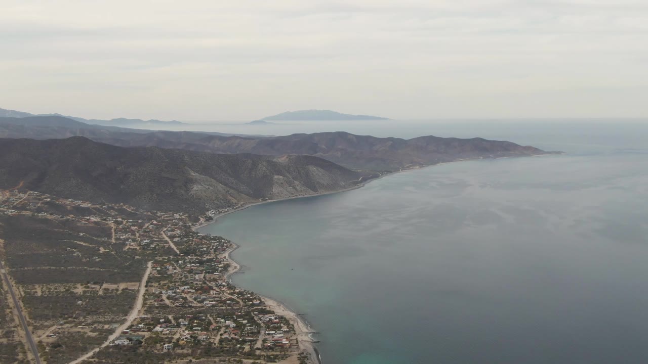 Drone frame over Los Barriles with pale roads and scattered buildings below the hillside, the shoreline bending into blue water, a level horizon, and no visible people.