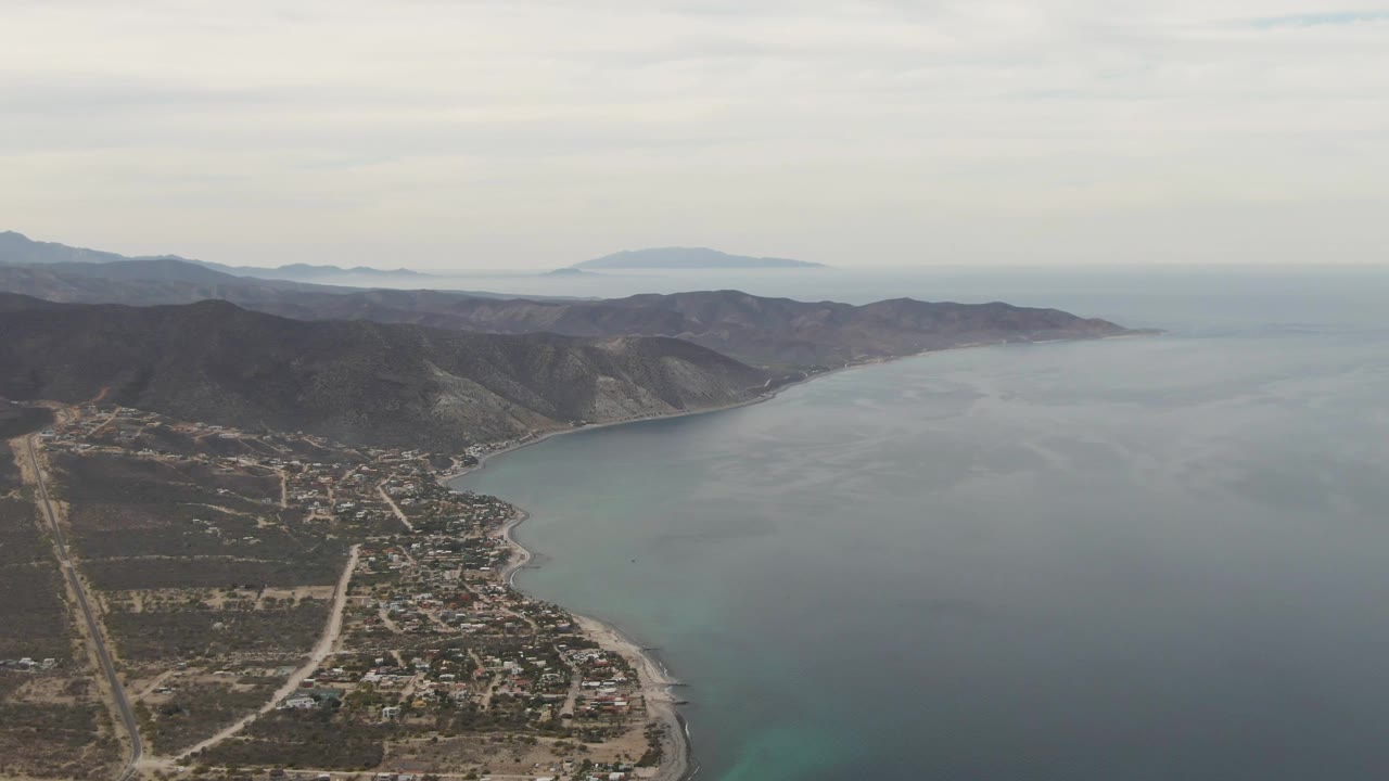 Wide aerial view of Los Barriles with the town near the lower center, dry hills stretching along the coast, calm blue sea to the right, distant land on the horizon, and no visible people.