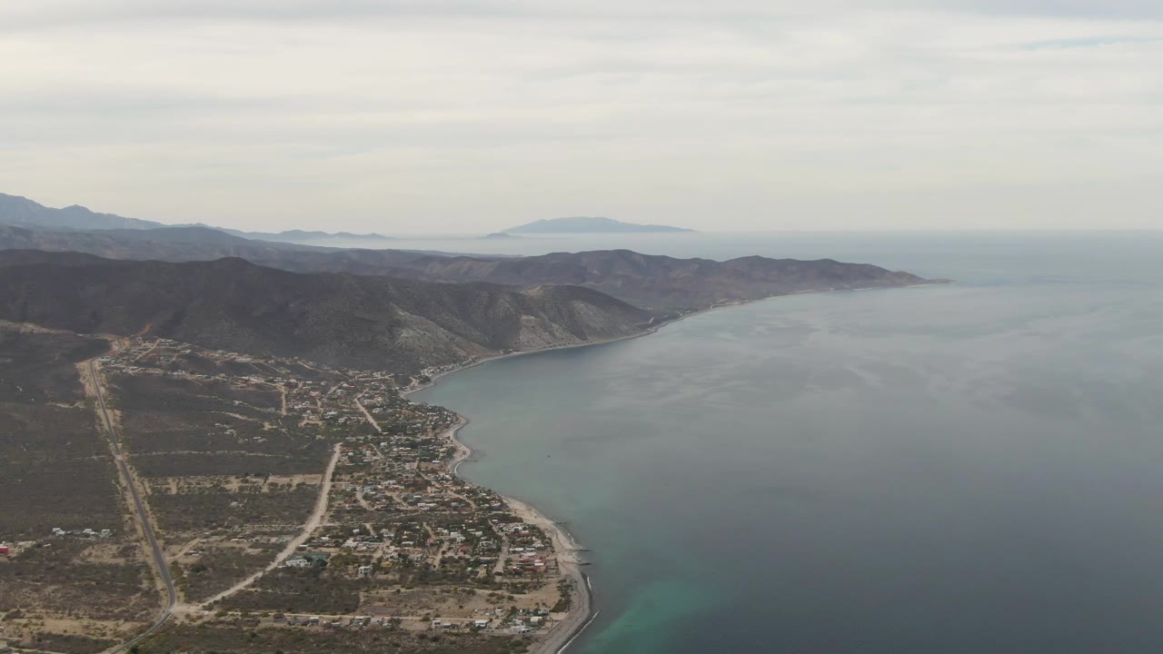 Aerial view looking along a curved coastline with roads, homes, hills, and sea.