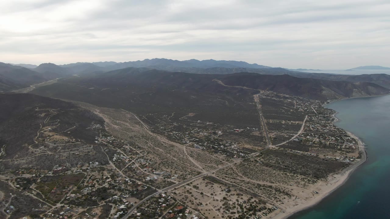 Aerial view over Los Barriles with blue-green sea on the right, sandy shoreline below, dry roads and homes across the coast, dark mountain ridges in the distance, and no visible people.