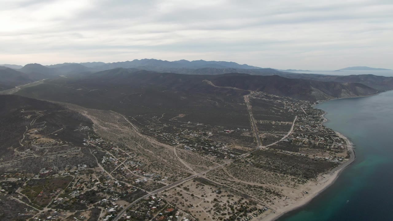 Wide aerial frame of Los Barriles showing a pale sandy beach curving along the bay, a grid of roads and houses inland, blue water to the right, layered mountains beyond, and no visible people.