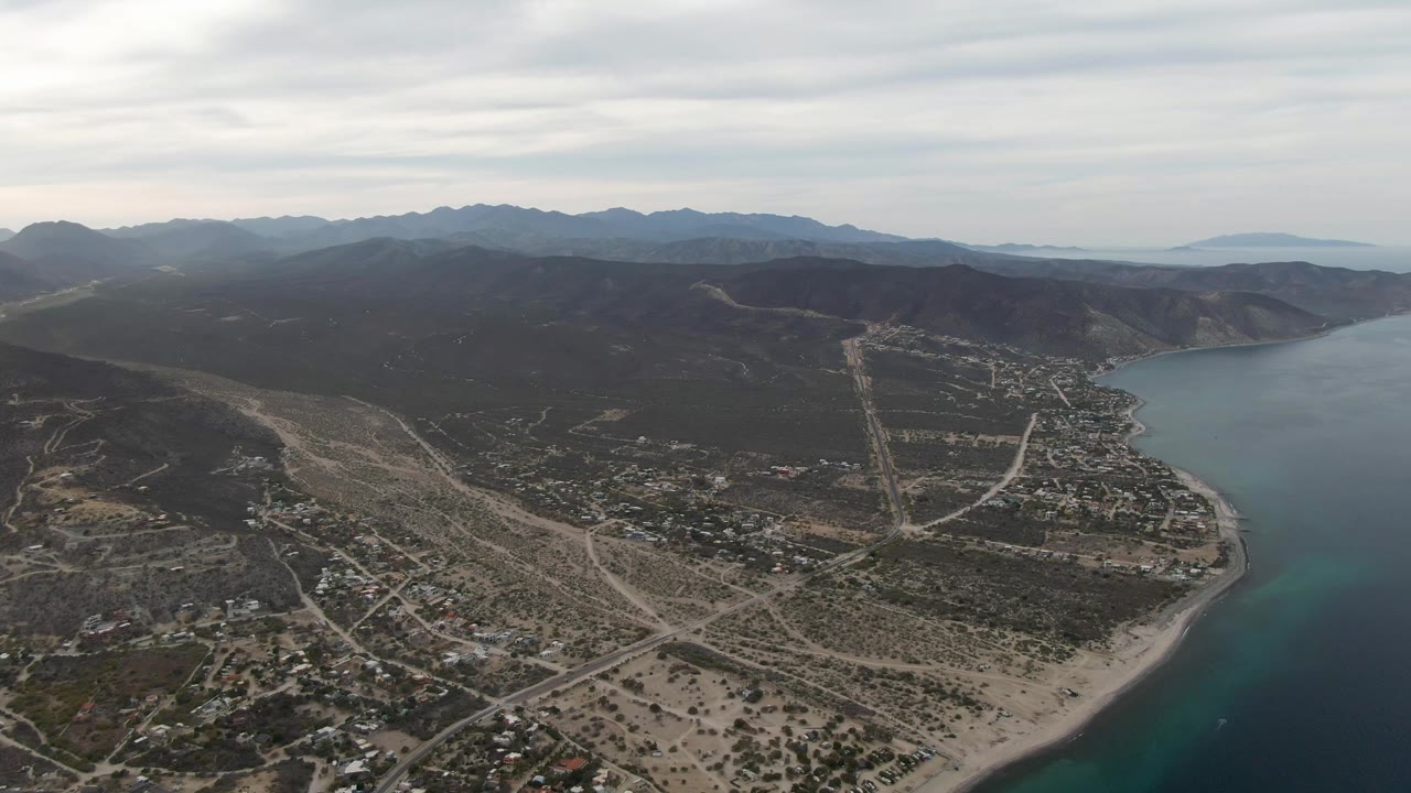 Drone-height view of Los Barriles with turquoise water along the lower right edge, sandy coastal lots and roads in the foreground, darker hills behind town, and no visible people.
