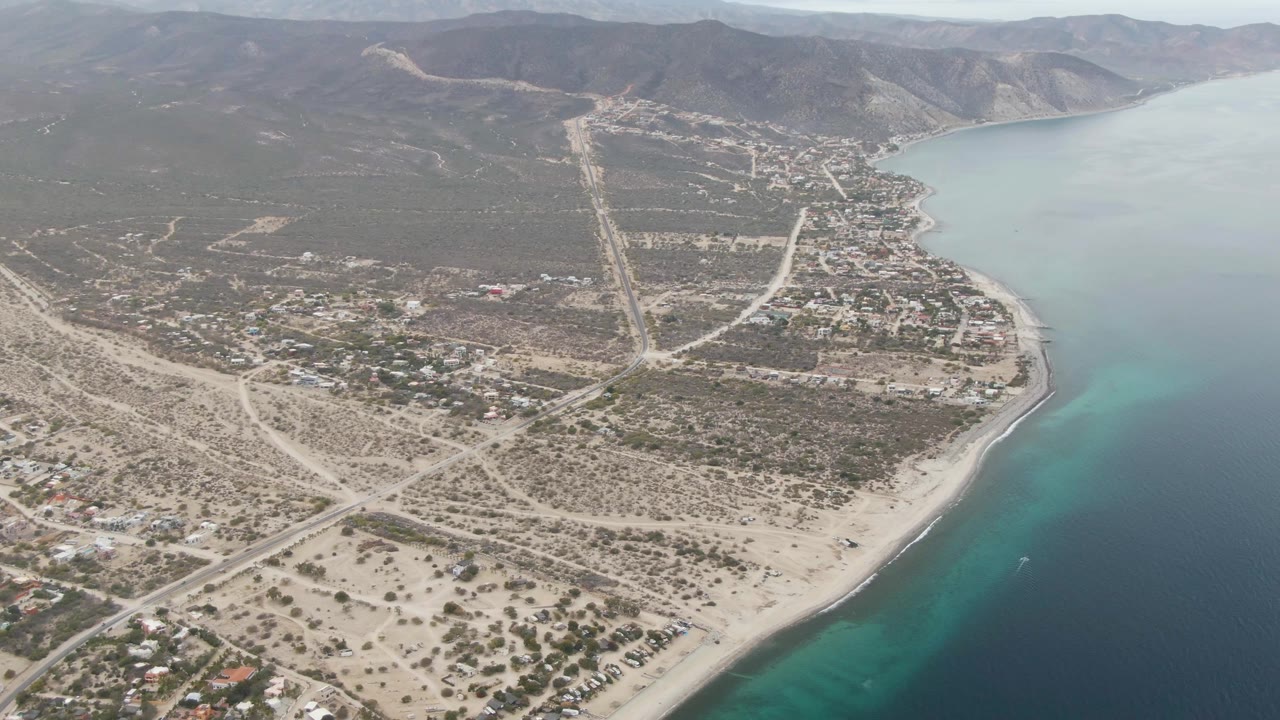 Aerial frame over Los Barriles, Baja California Sur, showing sandy shoreline, turquoise shallows, scattered buildings, pale roads, arid hills, and no visible people.