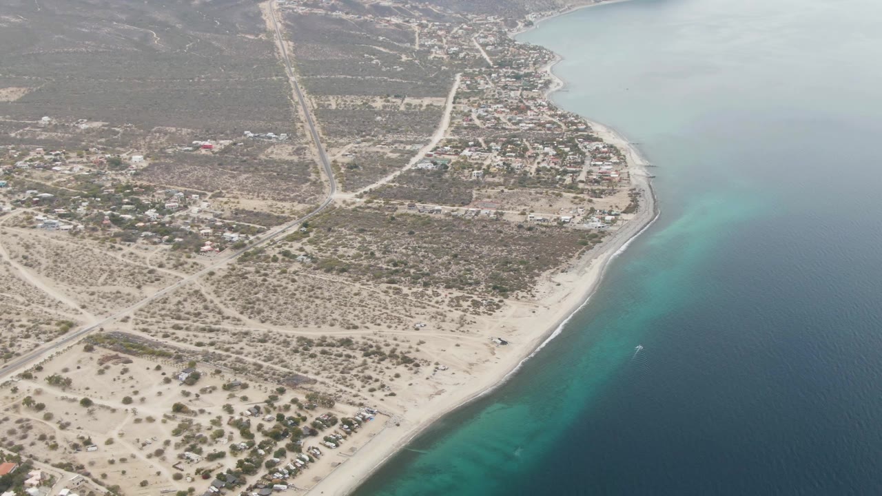 Oblique drone view over Los Barriles showing the shoreline curve, pale beach lots, inland roads, sparse vegetation, distant mountains, and no visible people.