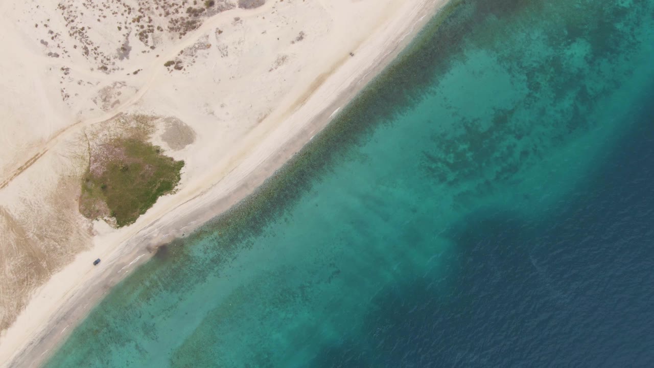 Aerial view in Los Barriles, Baja California Sur with the vehicle slightly farther along the sandy road, scrub-covered ground at left, bright beach, turquoise water, and no visible people.