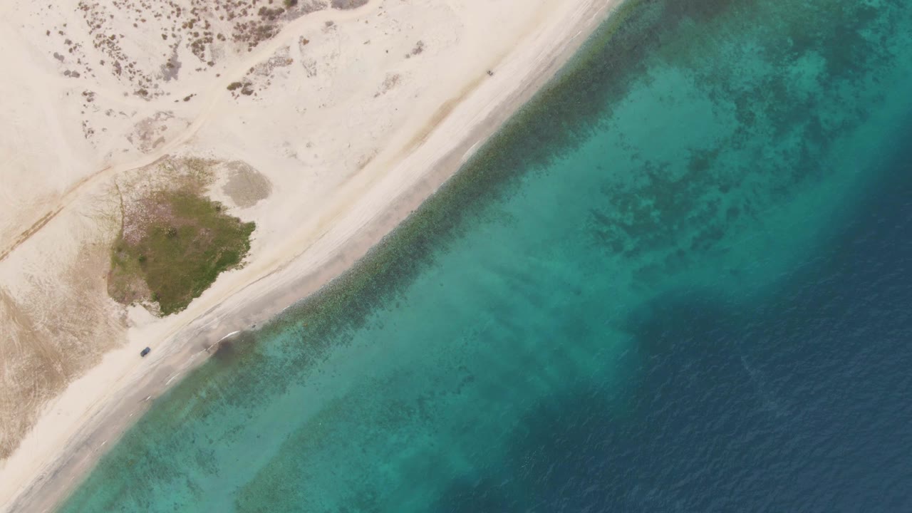 Drone frame from Los Barriles, Baja California Sur showing the vehicle on the diagonal sandy road, a rounded patch of vegetation inland, clear turquoise shallows, darker water offshore, and no visible people.