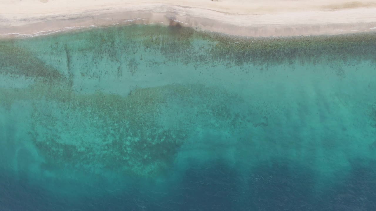 Top-down frame from Los Barriles showing a narrow pale beach, a light surf trace, green-blue shallows, and darker underwater patches spreading toward deeper water, with no visible people.