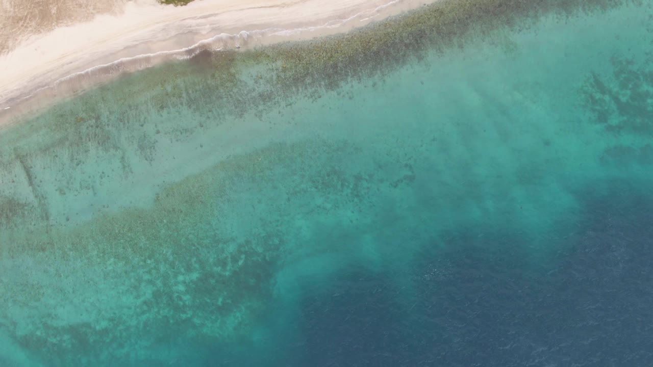 Overhead Los Barriles view with white sand at the upper edge, broad turquoise water in the middle, darker underwater formations, and no visible people.