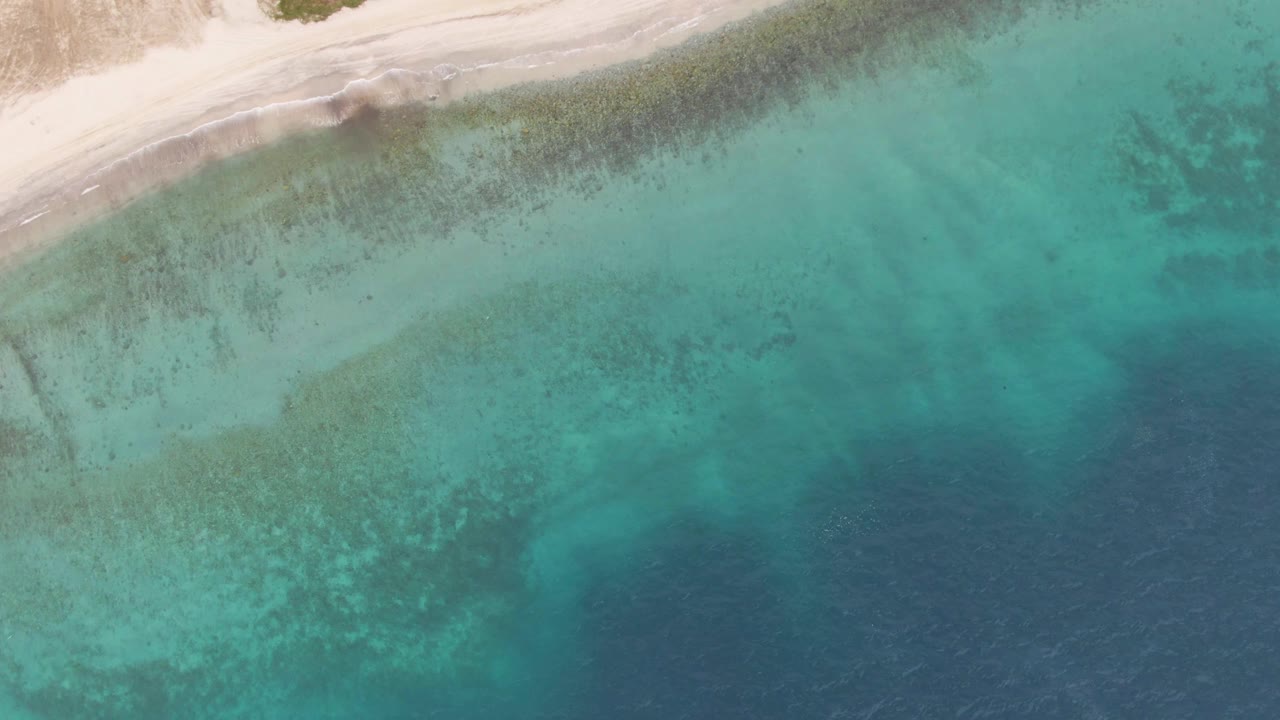 Drone-style overhead frame from Los Barriles with a diagonal white sand shoreline, faint wave foam, clear green-blue shallows, and no visible people.