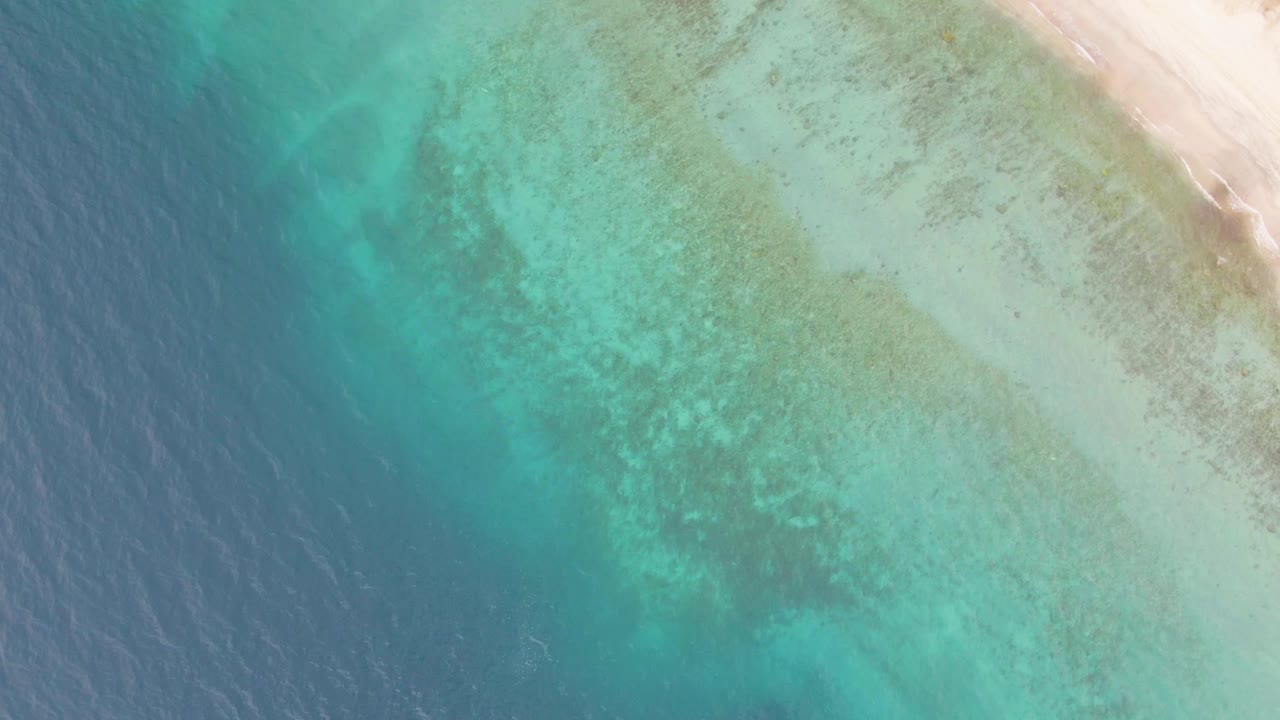 Aerial frame over Los Barriles showing teal water, darker reef-like seabed patches, and a narrow sandy shoreline at the upper right, with no visible people.