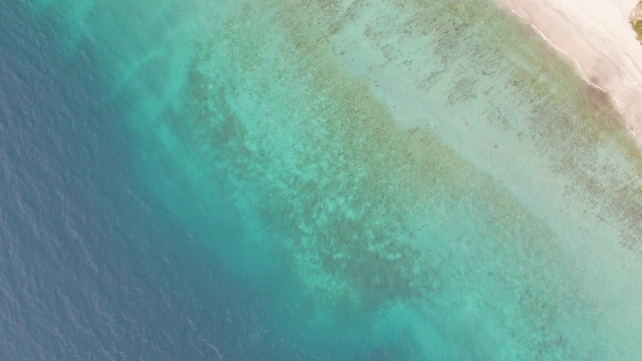 Overhead Los Barriles coastline view with deeper blue water on the left, pale turquoise shallows across the center, and sand along the top right, with no visible people.