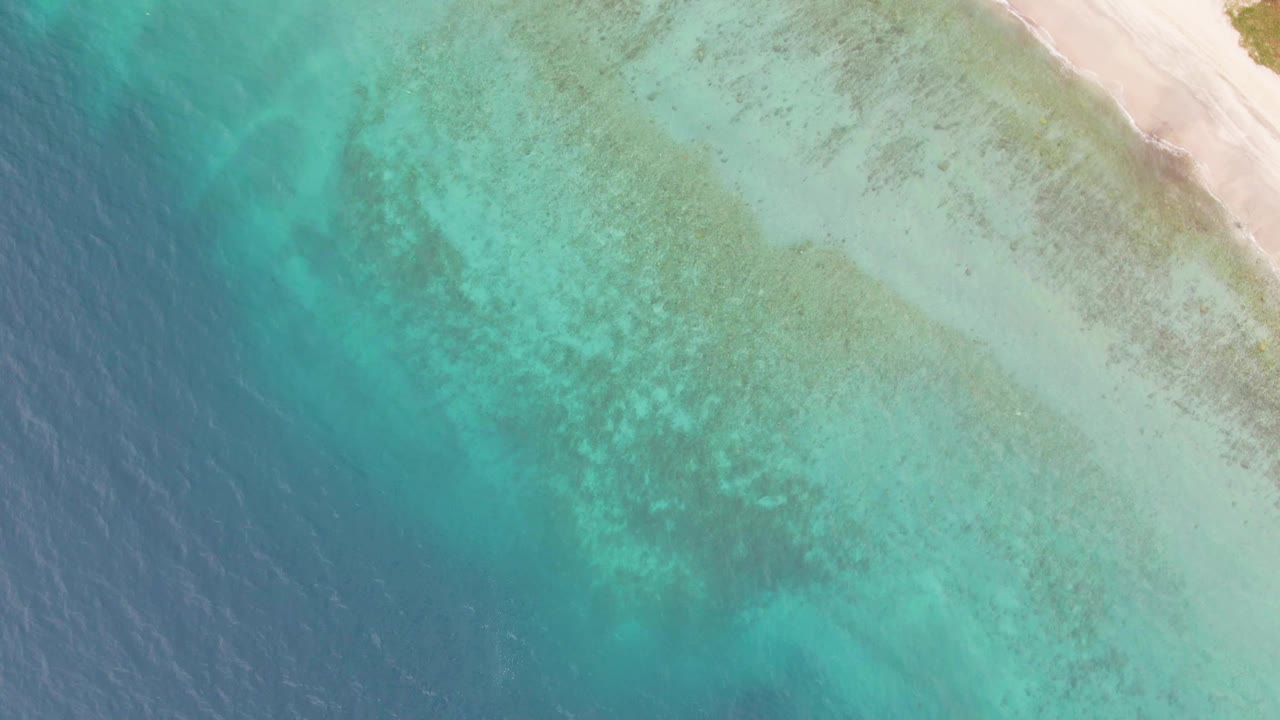 Aerial view at Los Barriles with broad turquoise shallows filling the frame, deeper water at left, and a pale sand strip along the upper right, with no visible people.