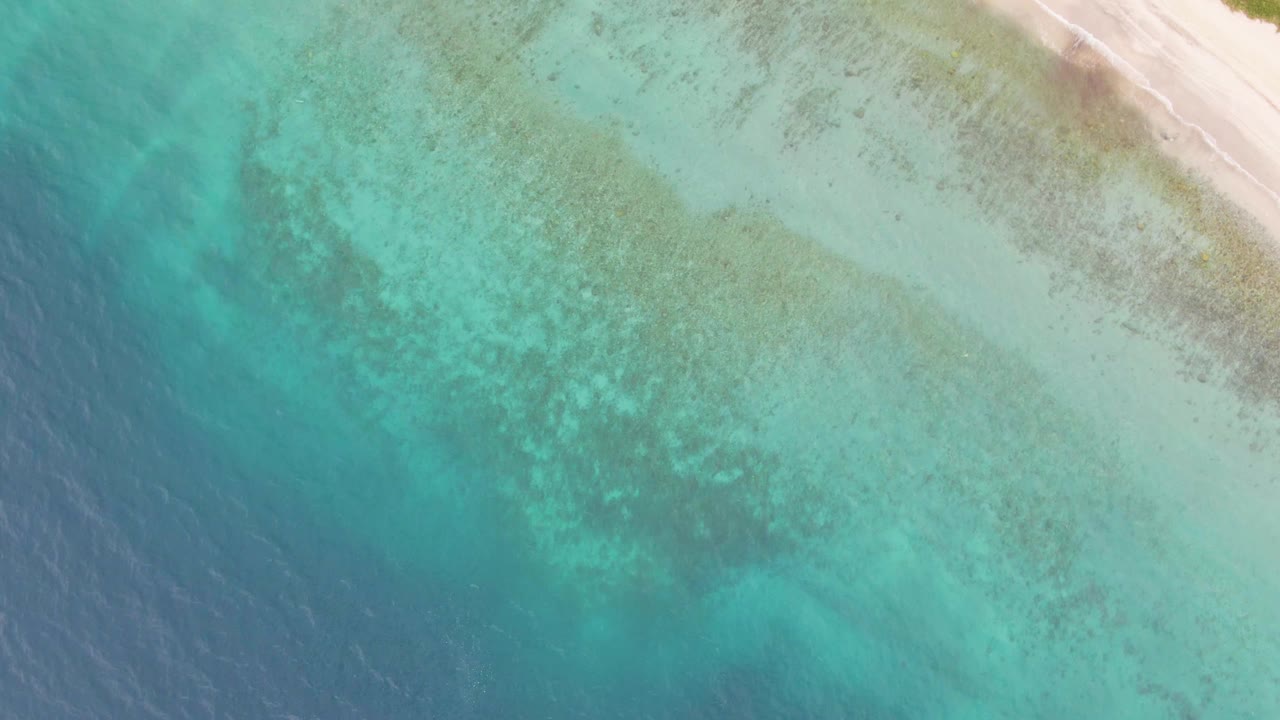Aerial view over Los Barriles showing pale sand at the upper right, shallow turquoise water with seabed texture, darker blue water at lower left, and no visible people.