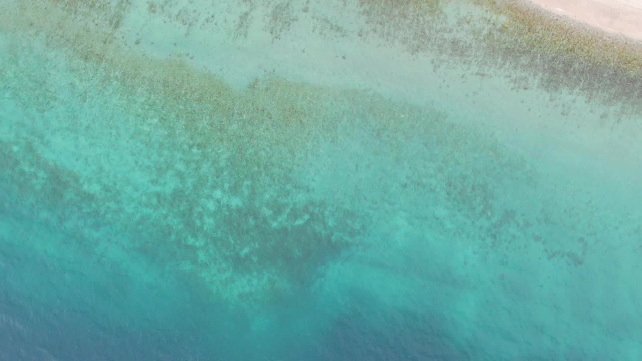 Aerial frame over Los Barriles where blue-green shallow water dominates the view, darker reef-like patches sit below the surface, and a thin sandy edge remains visible at the top, with no visible people.