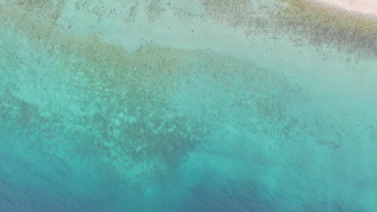 Aerial frame over Los Barriles with pale sand running across the upper frame, a light turquoise nearshore band, and darker coral-like seabed texture below, with no visible people.