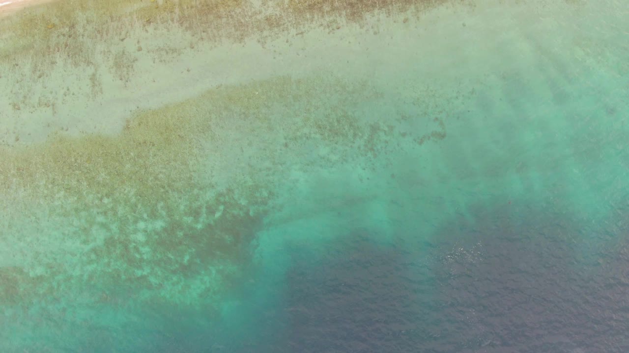 Straight-down aerial over Los Barriles, Mexico showing clear turquoise shallows, irregular seabed patches, deeper blue water along the lower frame, and no visible people.