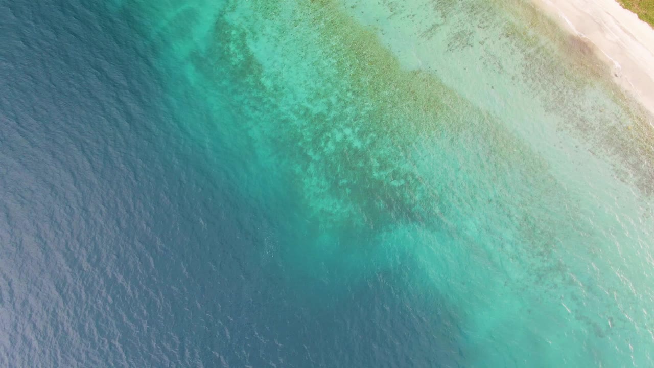 Aerial view over Los Barriles with the sandy shore near the top right, mottled seabed visible through aqua water, darker ocean to the left, and no visible people.