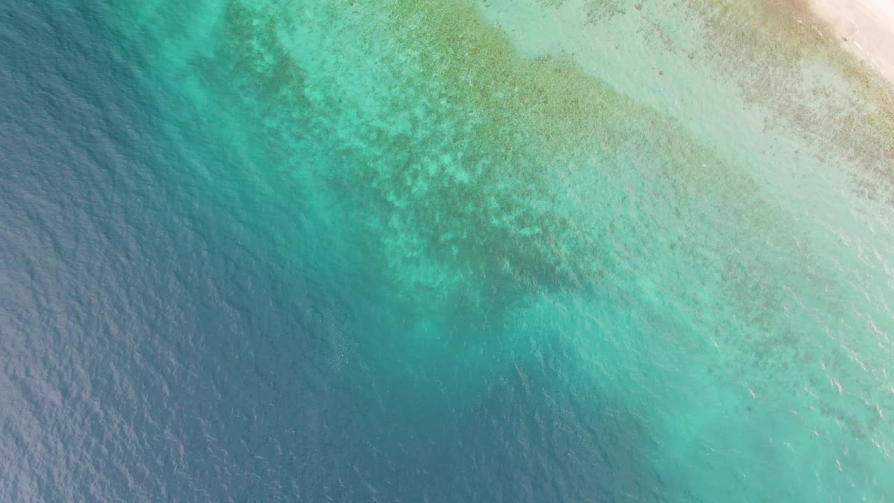 Mid-clip aerial frame over Los Barriles where the beach is mostly at the upper right edge, dark seabed patches sit under turquoise water, and no visible people appear.