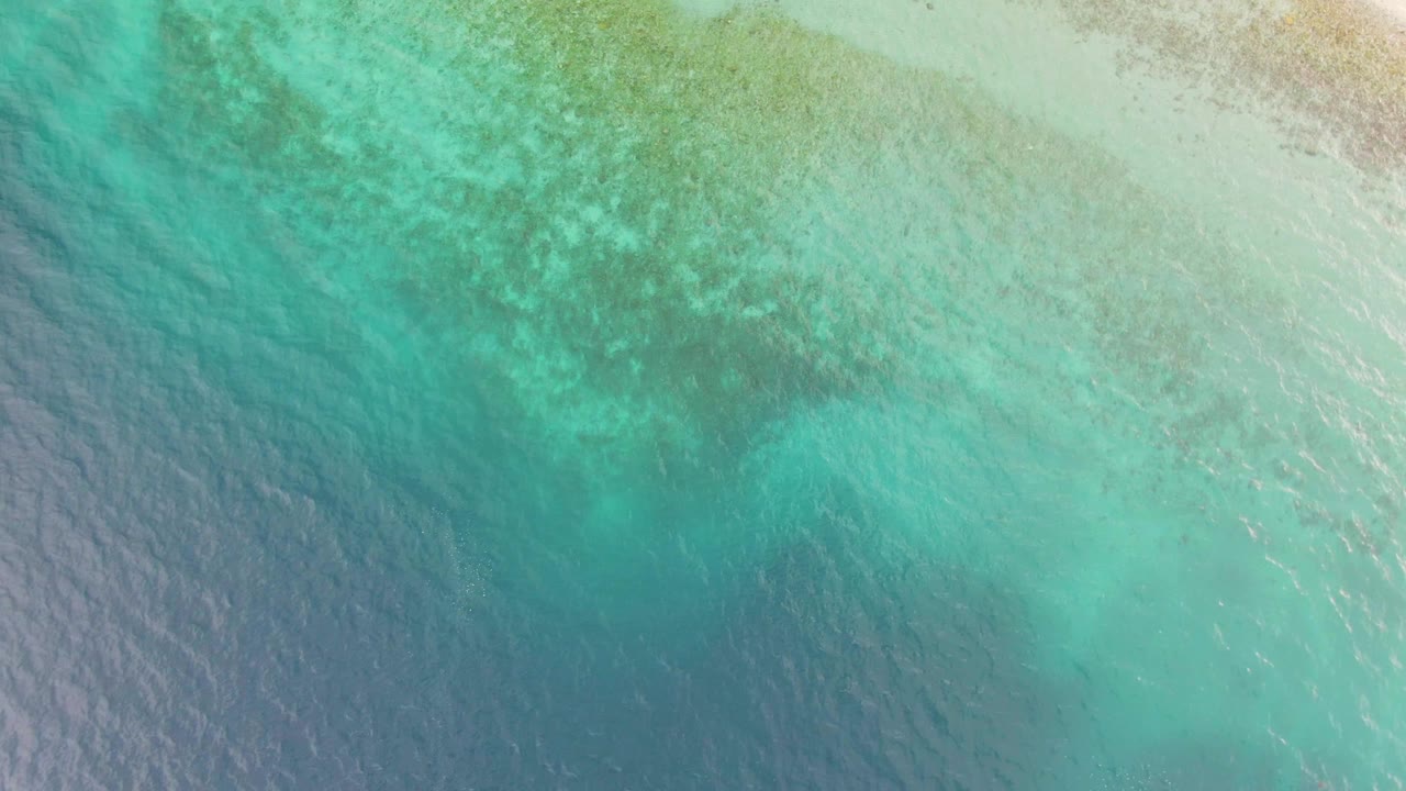 Clear blue-green water over a sandy seabed in Los Barriles, Mexico, with a broad darker reef patch near the upper frame and no visible people.