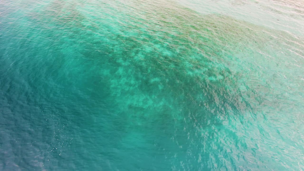Top-down view of turquoise sea in Los Barriles, Baja California Sur, where darker submerged areas sit below a lightly rippled surface, with no visible people.