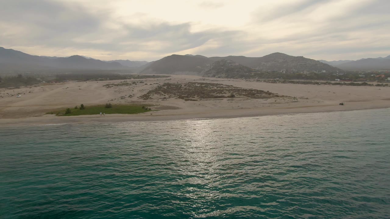 Opening frame of Los Barriles coastline with rippled turquoise water in the foreground, a pale sandy beach beyond it, hazy mountains in the distance, and no visible people.
