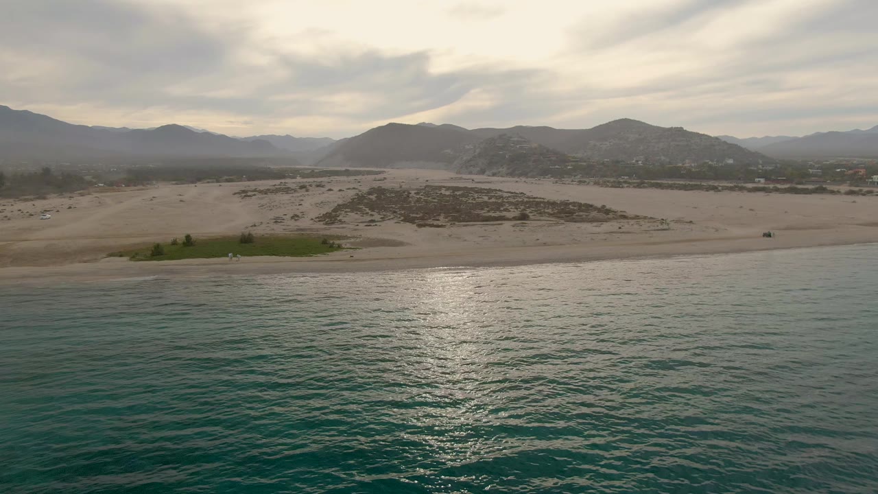 Los Barriles shoreline seen from above the water, with a brighter vertical reflection on the ocean surface, pale sand across the midground, mountain ridges behind, and no visible people.