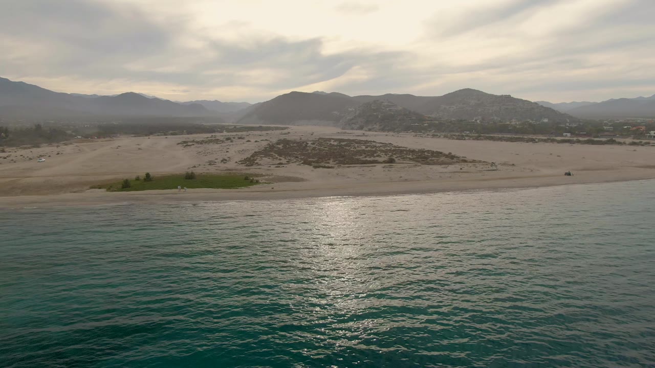 Aerial view over the Los Barriles coast with textured turquoise water across the lower frame, a mostly empty sandy beach, scattered low vegetation, distant mountains, and no visible people.