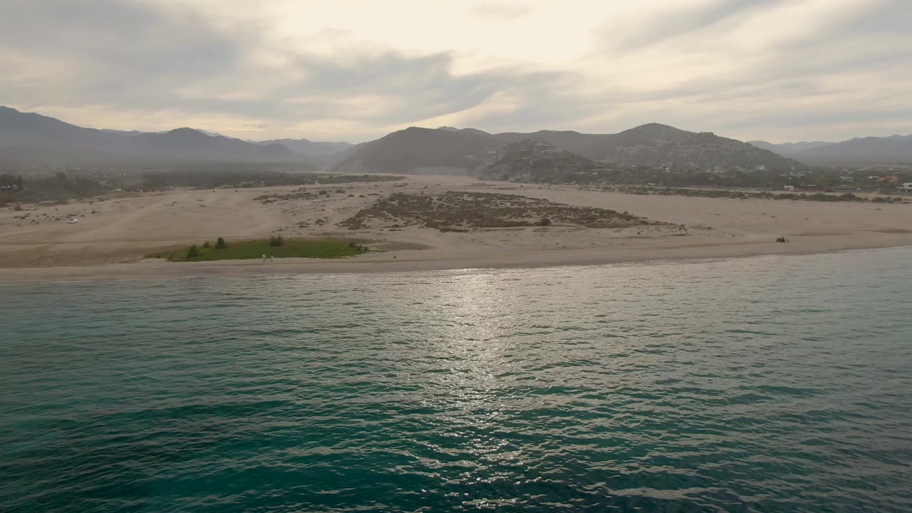 Ocean ripples and sun reflection below a sandy beach with mountains in haze.