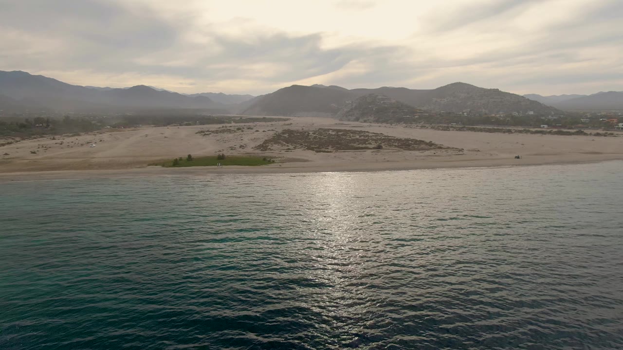 Calm ocean facing a sandy beach and distant mountains under cloudy light