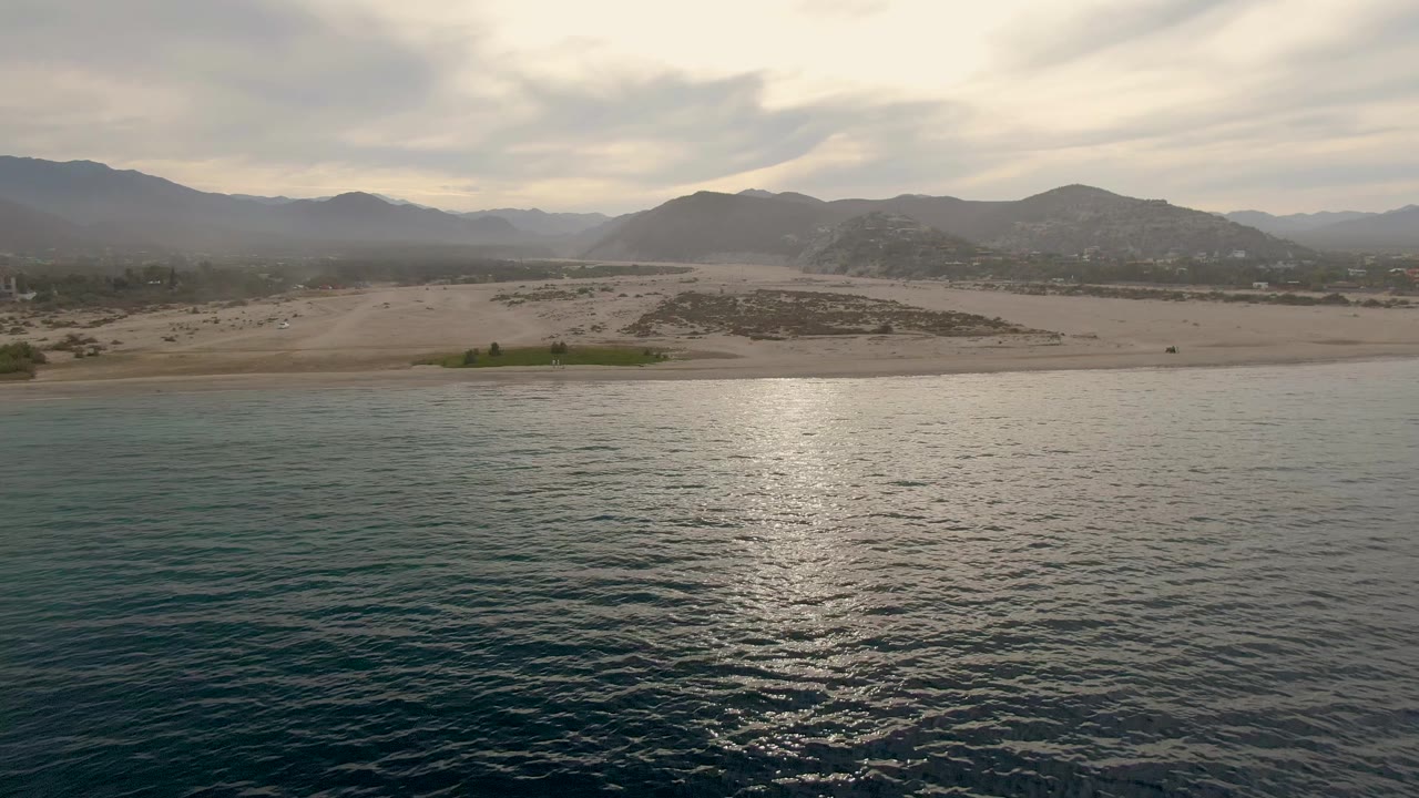 Third frame holds teal water in the foreground with an empty sandy beach, low shrubs, and hazy mountains around Los Barriles, with no visible people.