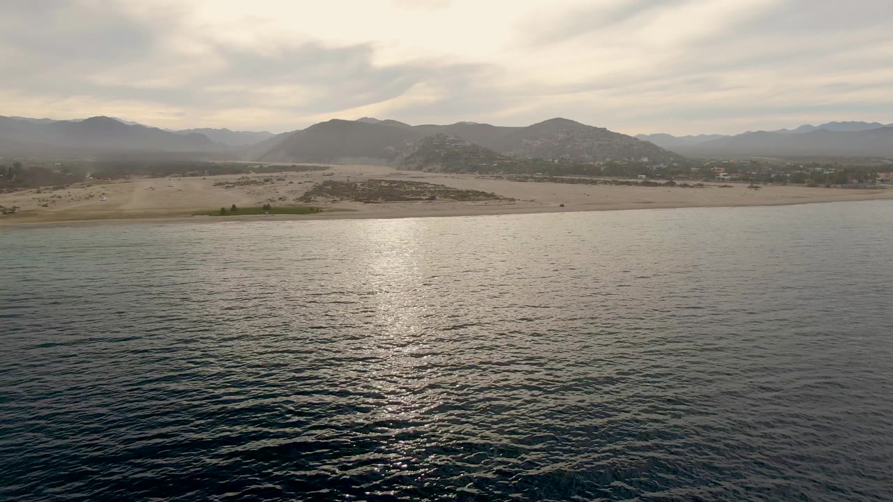 Opening aerial frame over Los Barriles with dark rippled ocean in front, a pale sandy shoreline, distant mountains, cloud-filtered light, and no visible people.