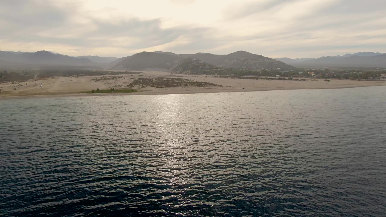 Third frame from the Los Barriles aerial view with textured ocean foreground, a narrow sandy beach, low coastal terrain, layered mountains, and no visible people.