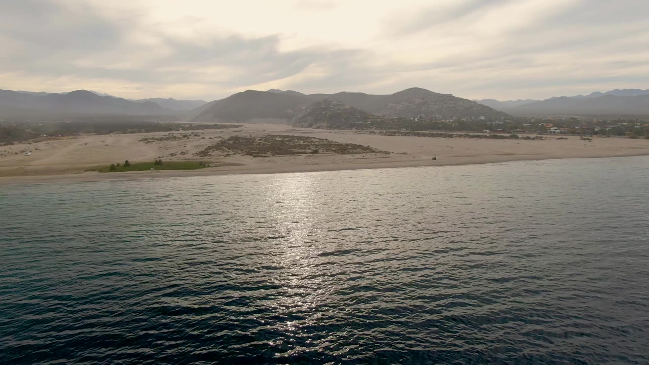 Second frame showing a brighter band of reflected light on the ocean, pale sand along the Los Barriles shoreline, distant mountains, cloud cover, and no visible people.