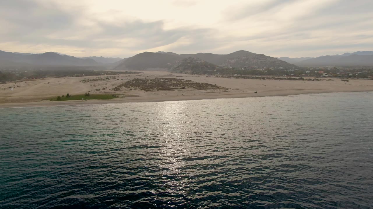 Third frame with darker teal and turquoise water in the foreground, the sandy Los Barriles beach crossing the midground, hazy mountains in back, layered clouds, and no visible people.