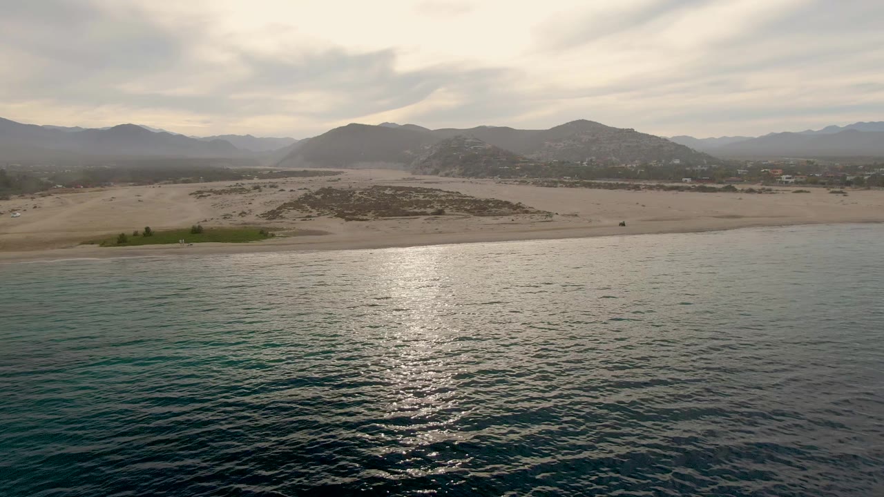 Fourth frame of the Los Barriles coast with calm water in front, pale sand and a small green patch near the shoreline, distant hills and mountains, muted clouds, and no visible people.