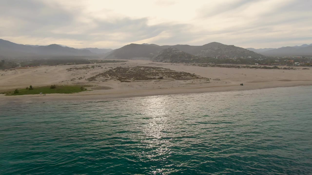 Aerial view of turquoise water, sandy beach, dunes, and distant mountains under clouds.