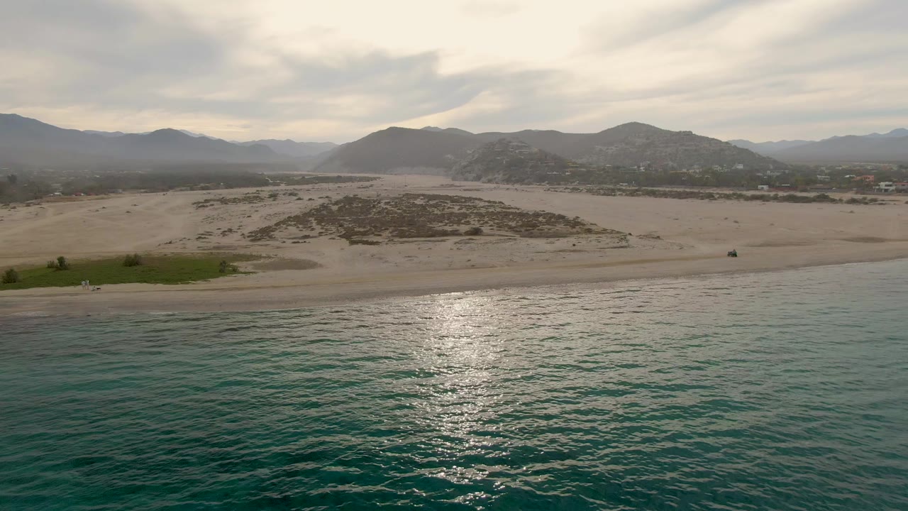 Los Barriles beach aerial with calm green-blue water in front, a narrow surf line, broad sandy dunes, scattered scrub, hazy mountains, layered clouds, and no visible people.