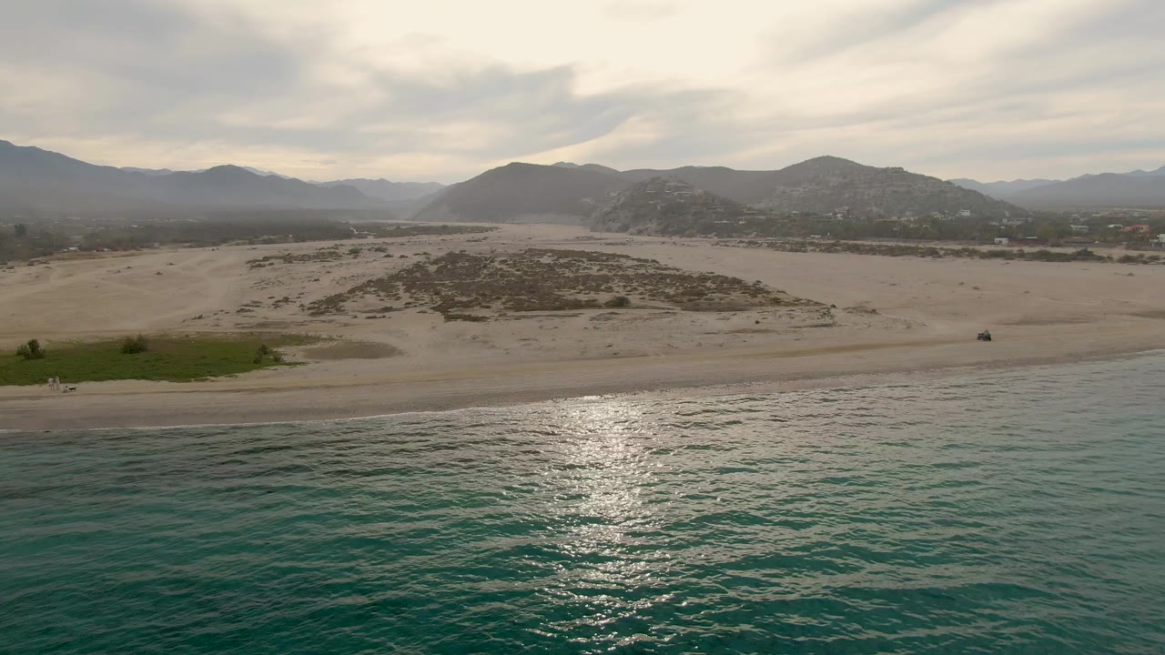 Aerial frame from Los Barriles showing empty shoreline, turquoise water with bright glare, a green patch near the sand, low dunes, distant mountain ridges, cloudy sky, and no visible people.
