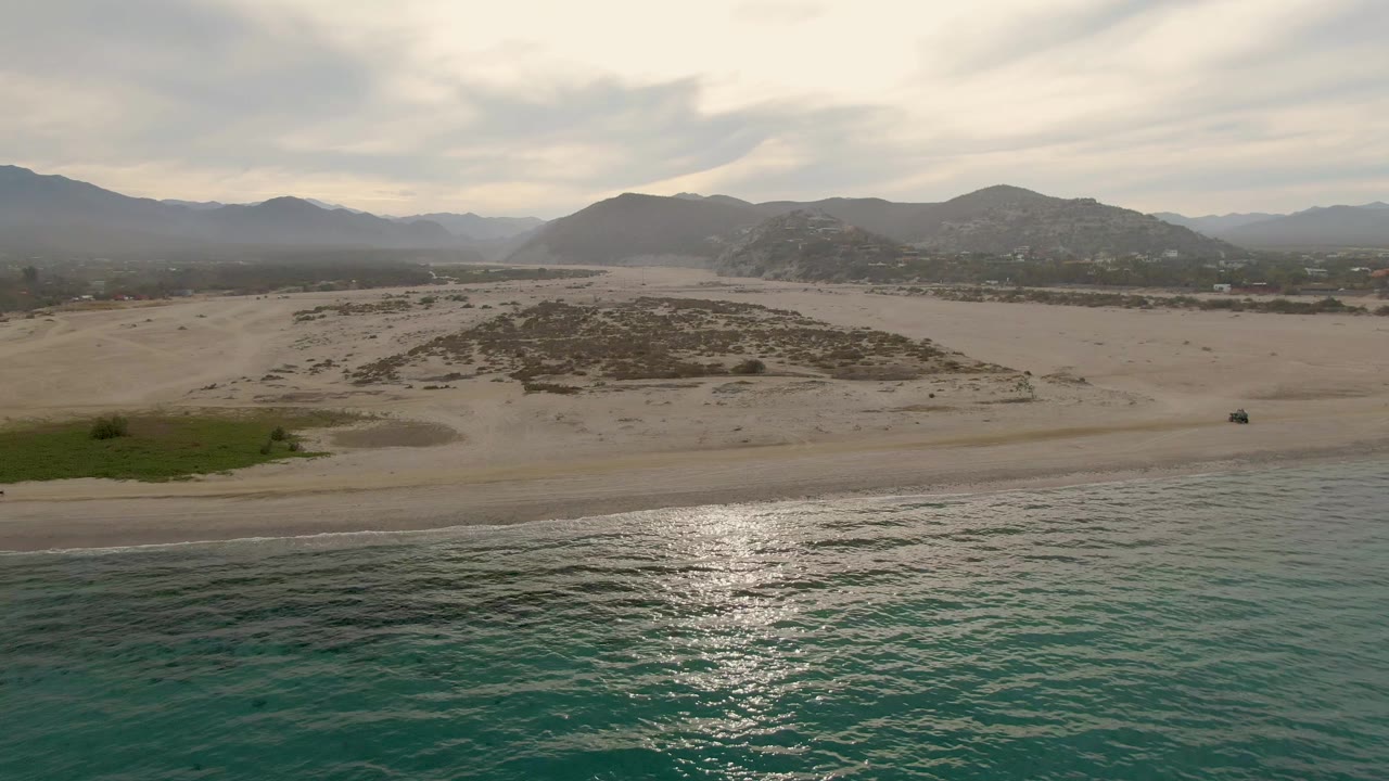 Los Barriles coastal aerial with ocean glare along the lower frame, pale beach and low dunes across the middle, green vegetation at the left edge, distant mountains, and no visible people.