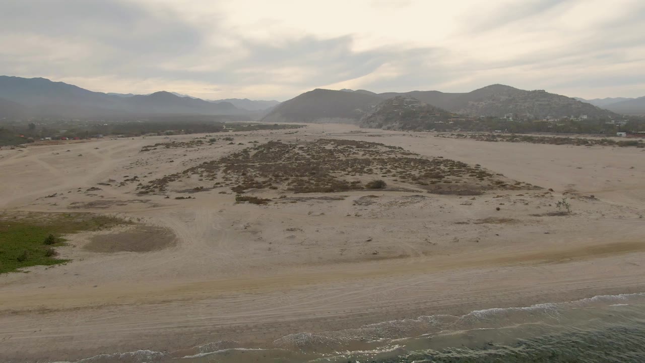 Aerial frame over sandy terrain near Los Barriles, Baja California Sur, with a strip of coastal water at the lower edge, scattered bushes, distant mountains, overcast sky, and no visible people.