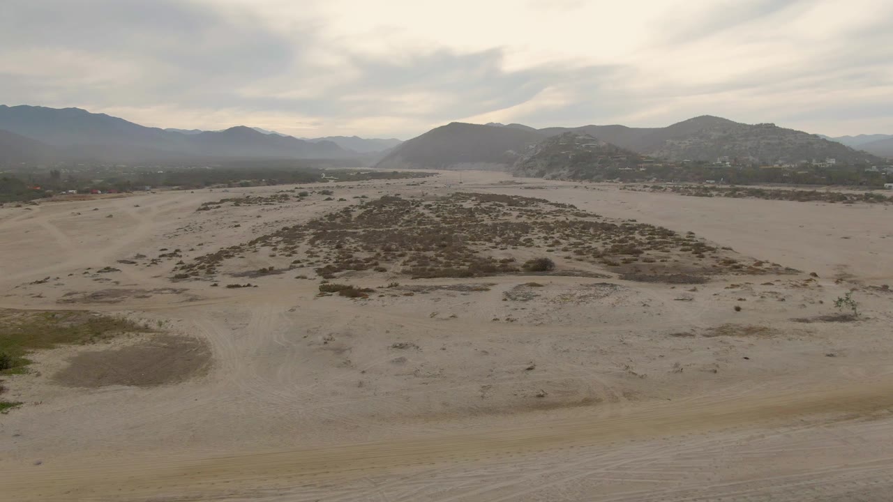 Wide sandy flat with low bushes, faint tracks, and mountains under cloud cover.